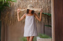 A model wearing a beige and white vertically striped sleeveless dress and a wide-brimmed straw hat, posing in front of a rustic thatched wall.